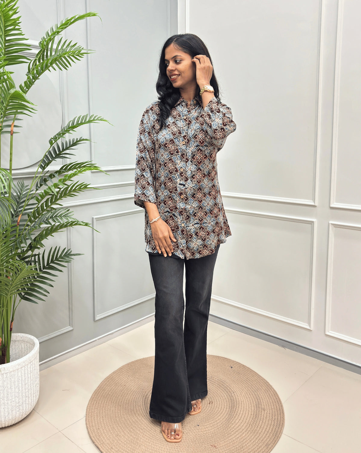 Black Women's Ajrakh Pure Modal Silk Shirt with a Floral print, black flared pants, standing on a round woven rug in front of a white paneled wall with a potted plant on the left.