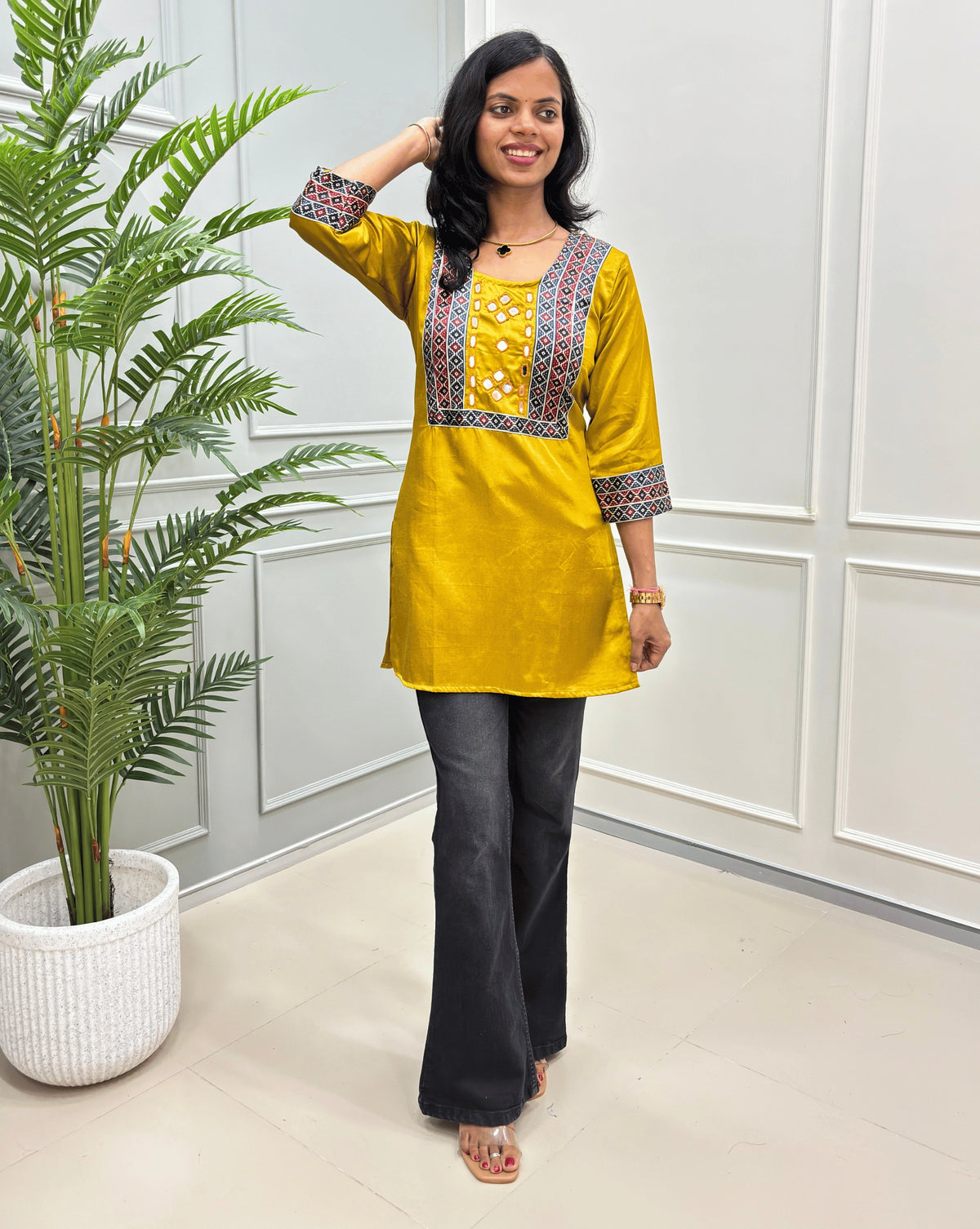  Mustard yellow women's mashru silk short kurti with colorful geometric embroidery on the chest and cuffs, paired with black flared pants, standing in a bright room with a potted plant.