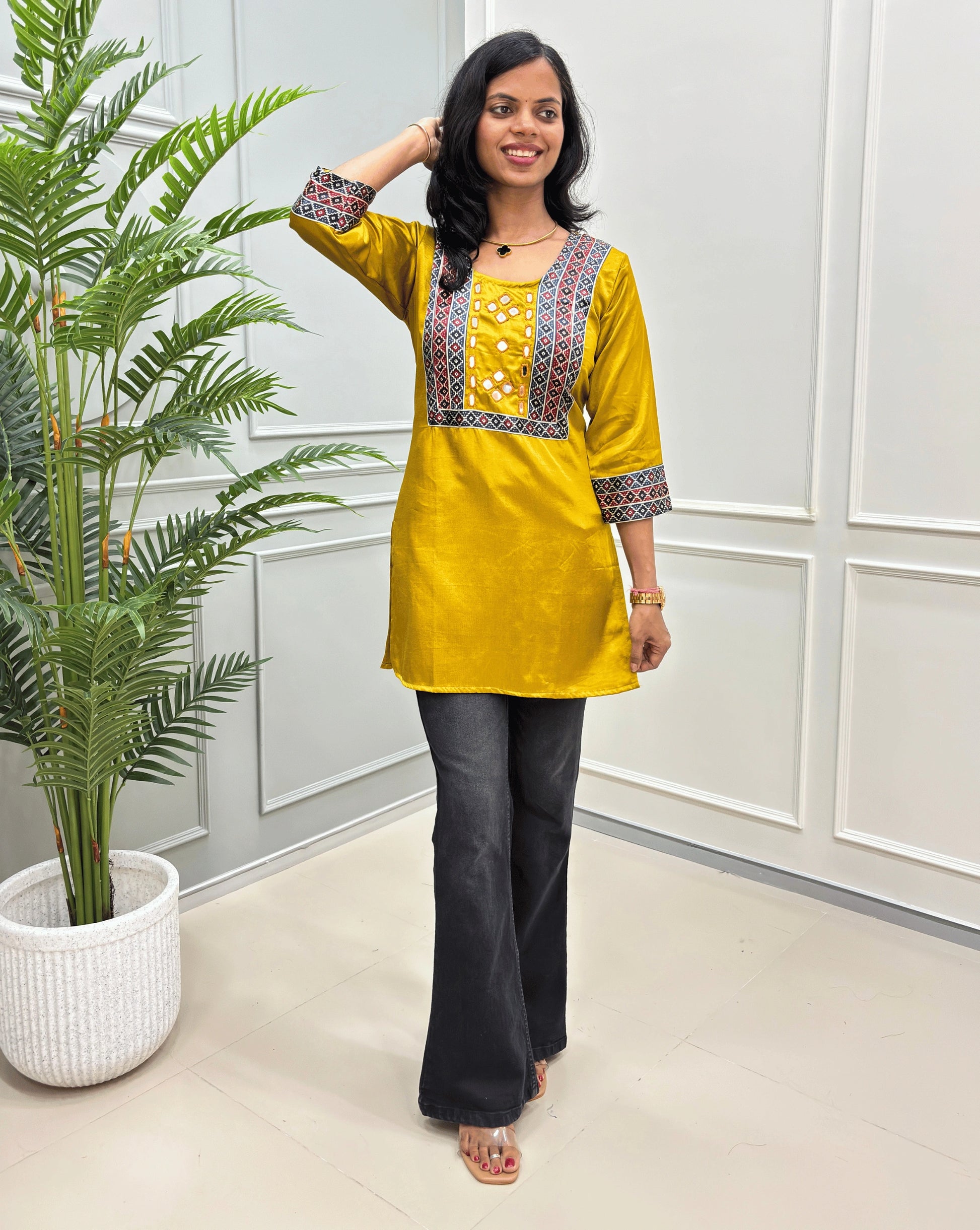  Mustard yellow women's mashru silk short kurti with colorful geometric embroidery on the chest and cuffs, paired with black flared pants, standing in a bright room with a potted plant.