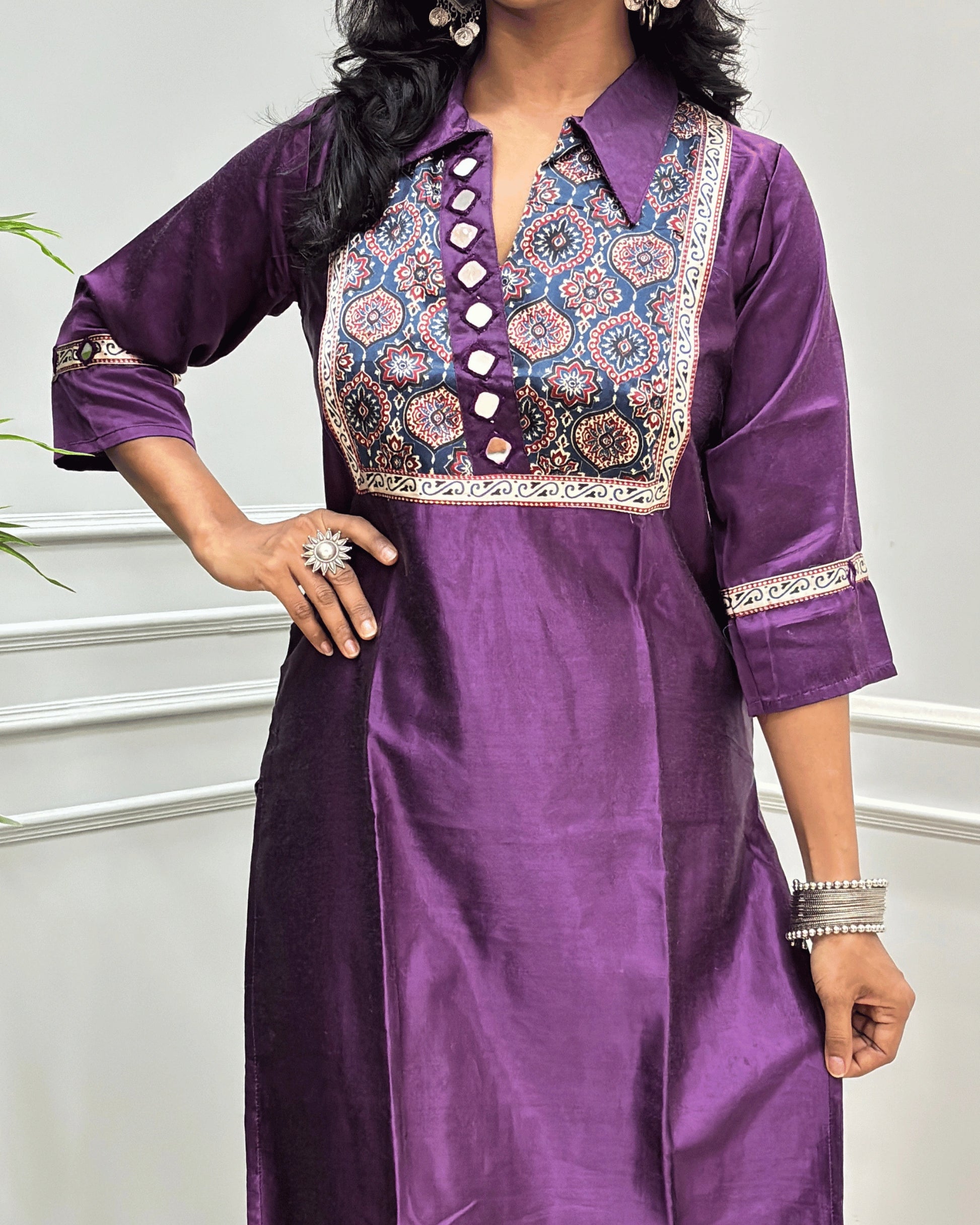 Close-up of Purple Ajrakh Mashru Silk Long-kurti with a heavily embroidered Badam-buta yoke and patterned sleeves, Hand-mirror Work on Front & Sleeves, Collar V- neck,  standing against a light gray paneled wall.