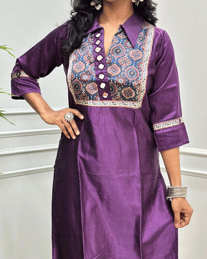 Close-up of Purple Ajrakh Mashru Silk Long-kurti with a heavily embroidered Badam-buta yoke and patterned sleeves, Hand-mirror Work on Front & Sleeves, Collar V- neck,  standing against a light gray paneled wall.