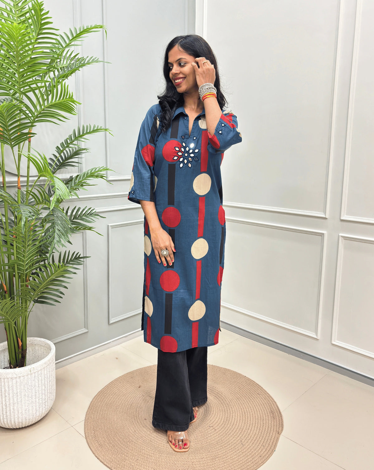 Deep-Blue Women's Ajrakh Vegetable dyed plant based Cotton Long-Kurti with a geometric pattern of Red, beige, and black circles, paired with black Flared pant, standing in a room with a potted plant and paneled walls.