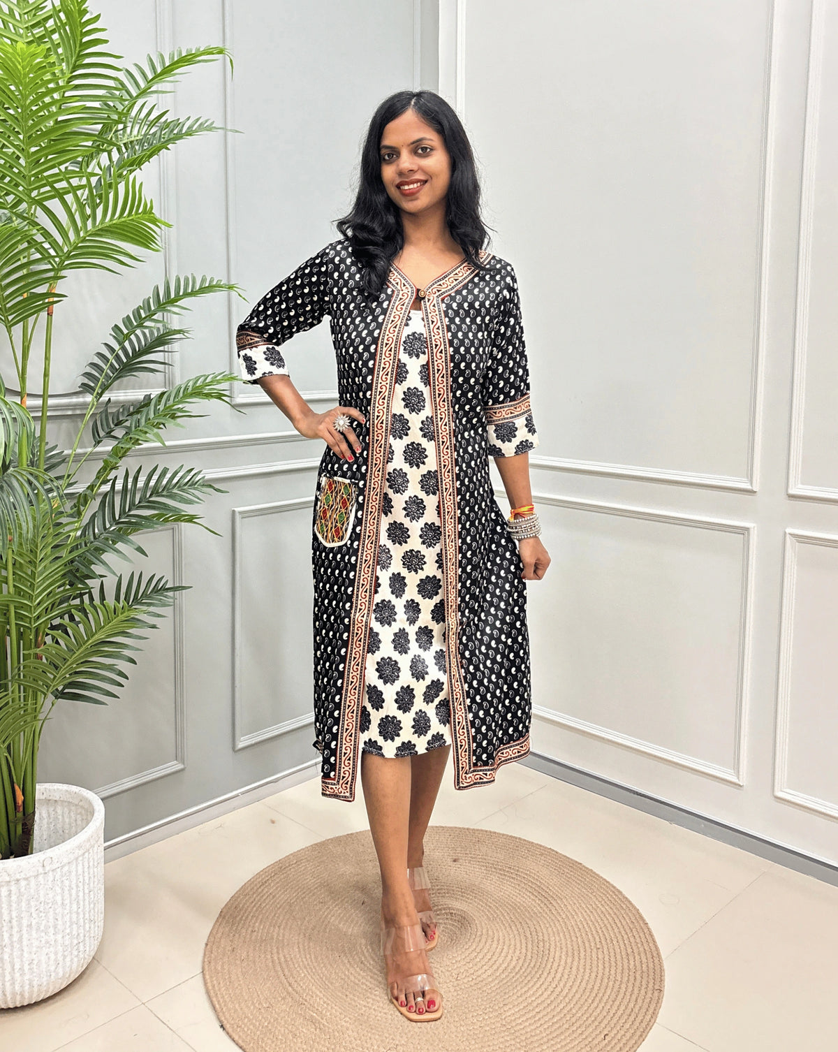Black women's Ajrakh pure Mashru silk Long one-peice with Black Shrug attach with White inner consist floral print wiith black colour , standing in a decorated room with a potted plant and paneled wall