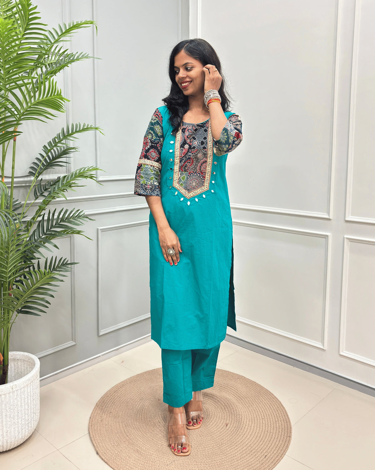 Aqua Blue women's Ajrakh Vegetable dyed plant based Cotton CoOrd set with Multi-colour ajrakh Print detailed embroidered on neckline & Sleevess with beige border on work ,  paired with matching teal pants, standing on a round woven mat in a bright room with a potted plant.