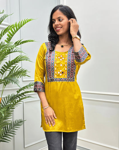 Close-up of handcrafted mirror work on yellow Ajrakh cotton kurti with colorful geometric embroidery on the neckline and ajrakh print cuffs, paired with dark gray pants, standing in front of a white paneled wall with a green plant on the left. 