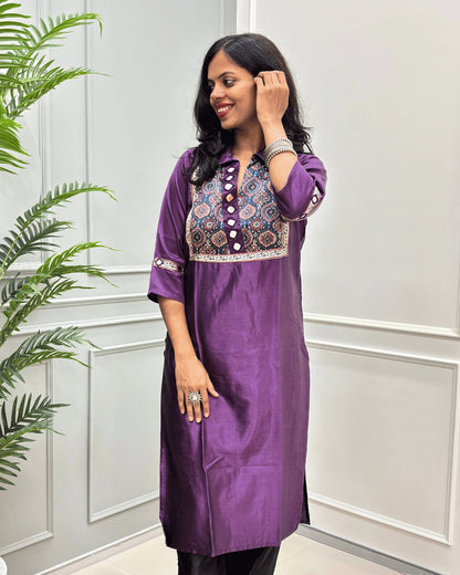 Front view of Purple women's Ajrakh Mashru Silk with three-quarter Sleeves Featured with detailed multicolor embroidered yoke and patterned sleeve cuffs with Badam Buta print , standing in front of a white paneled wall with a green plant on the left.