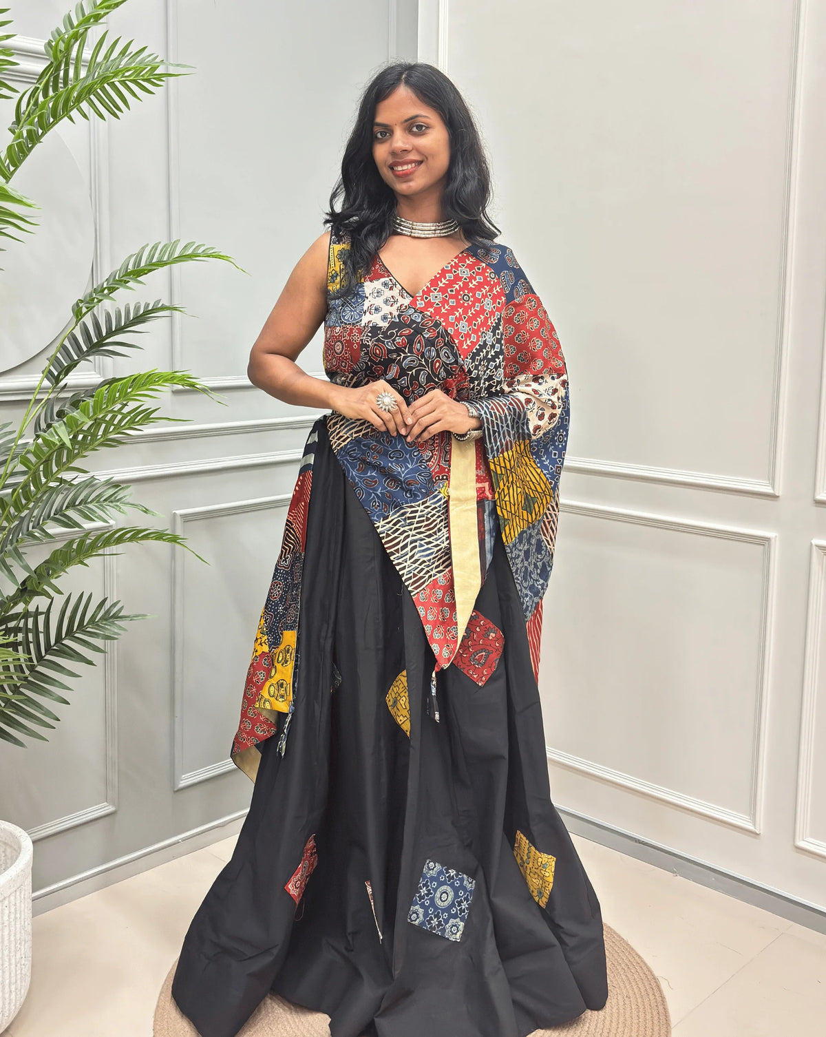 Front-view of Black Women's Ajrakh pure Cotton Skirt Set with a beautiful Tasals & Ajrakh Patch work along with multi-colour Printed blouse & Dupatta, Standing against a light paneled wall with a green plant on the left.