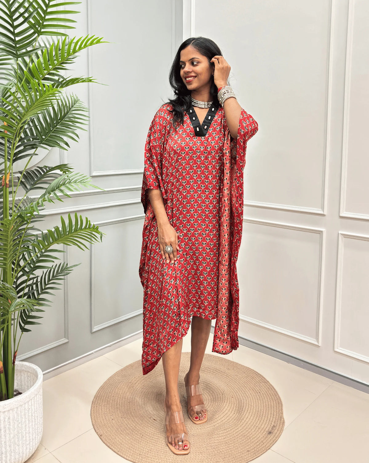 Red Women's Ajrakh Pure Modal Silk Knee Length Kaftan with a black Floral print , standing on a round woven rug in front of a white paneled wall next to a potted plant.