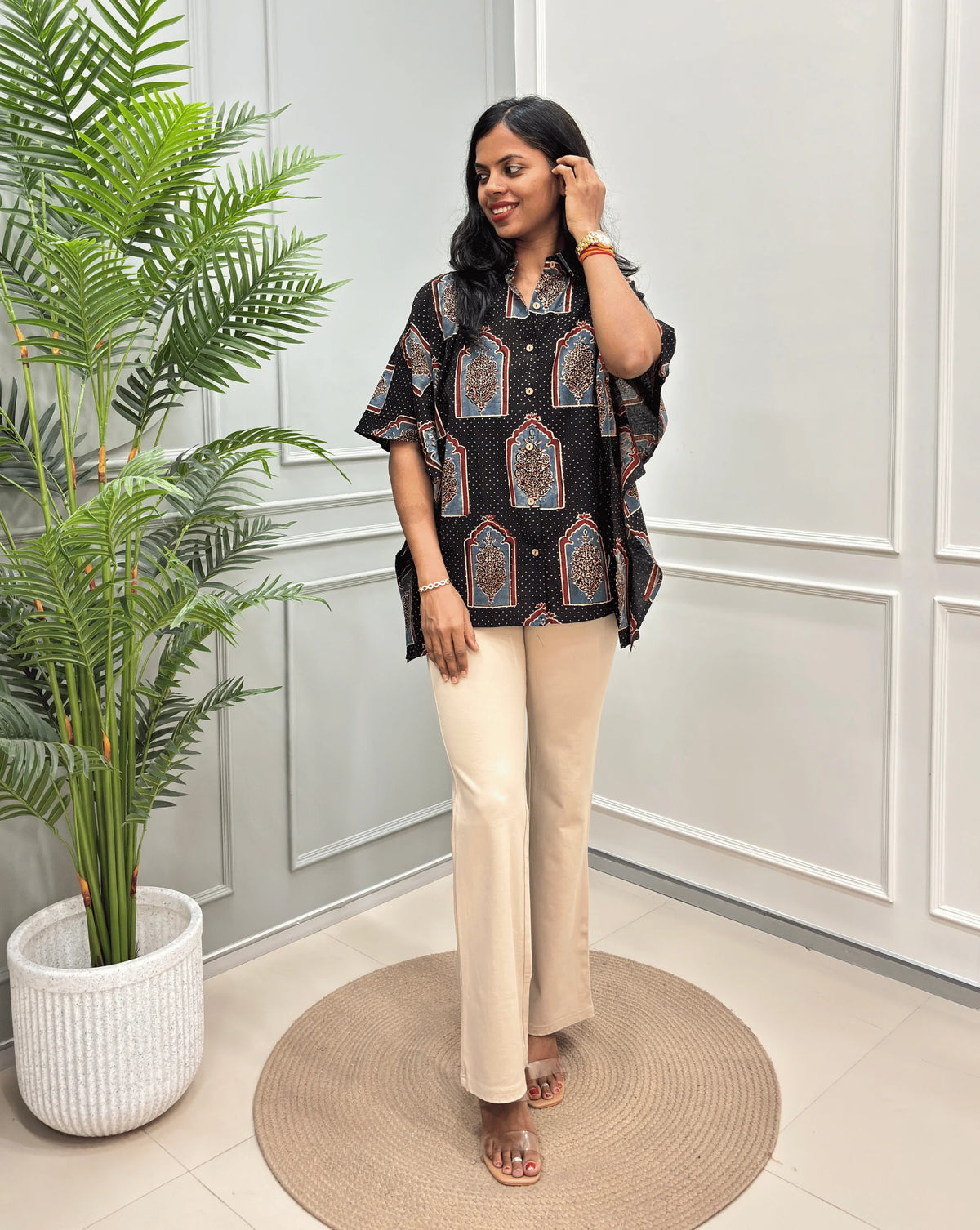 Black Women's Ajrakh Pure Cotton Short Kaftan Shirt with a Grey, Red & Black Floral Block Print, paired with beige flared pants, standing on a round woven rug in front of a white paneled wall with a potted plant.