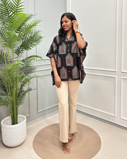 Black Women's Ajrakh Pure Cotton Short Kaftan Shirt with a Grey, Red & Black Floral Block Print, paired with beige flared pants, standing on a round woven rug in front of a white paneled wall with a potted plant.