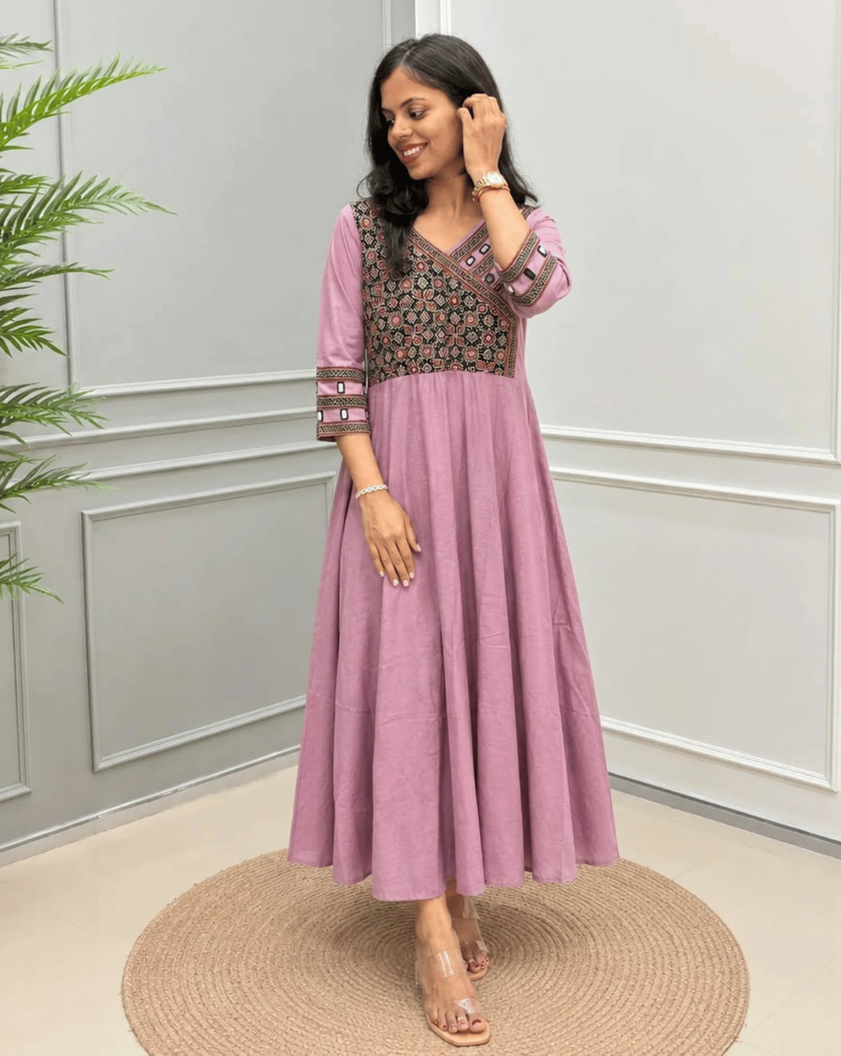Onion Pink  Women's Ajrakh Pure Cotton A-line Anarkali with a Black base & Red floral yoke patterned on Front & sleeve cuffs, standing on a round woven rug in front of a light gray paneled wall.