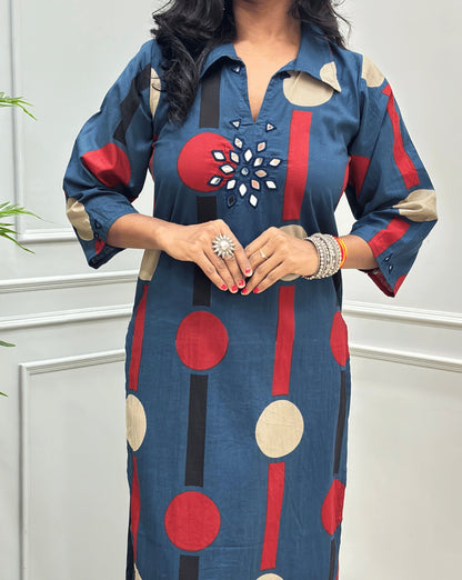 Close-up of Dark Blue Women's Ajrakh Vegetable dyed plant based Cotton Long-Kurti Featured Collar V-neck & Three-quarter Sleeves with a geometric pattern of Blue, beige, and black circles, Hand-mirror work on front in flower shape & Sleeves cuffs,  paired with black Flared pant, standing in a room with a potted plant and paneled walls.