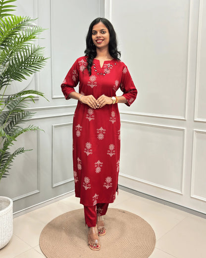Red Women's Ajrakh Vegetable Dyed Cotton Long Co-Ord set with white floral prints, along with Same Matching pant,  standing on a round woven mat in front of a white paneled wall with a potted plant on the left.