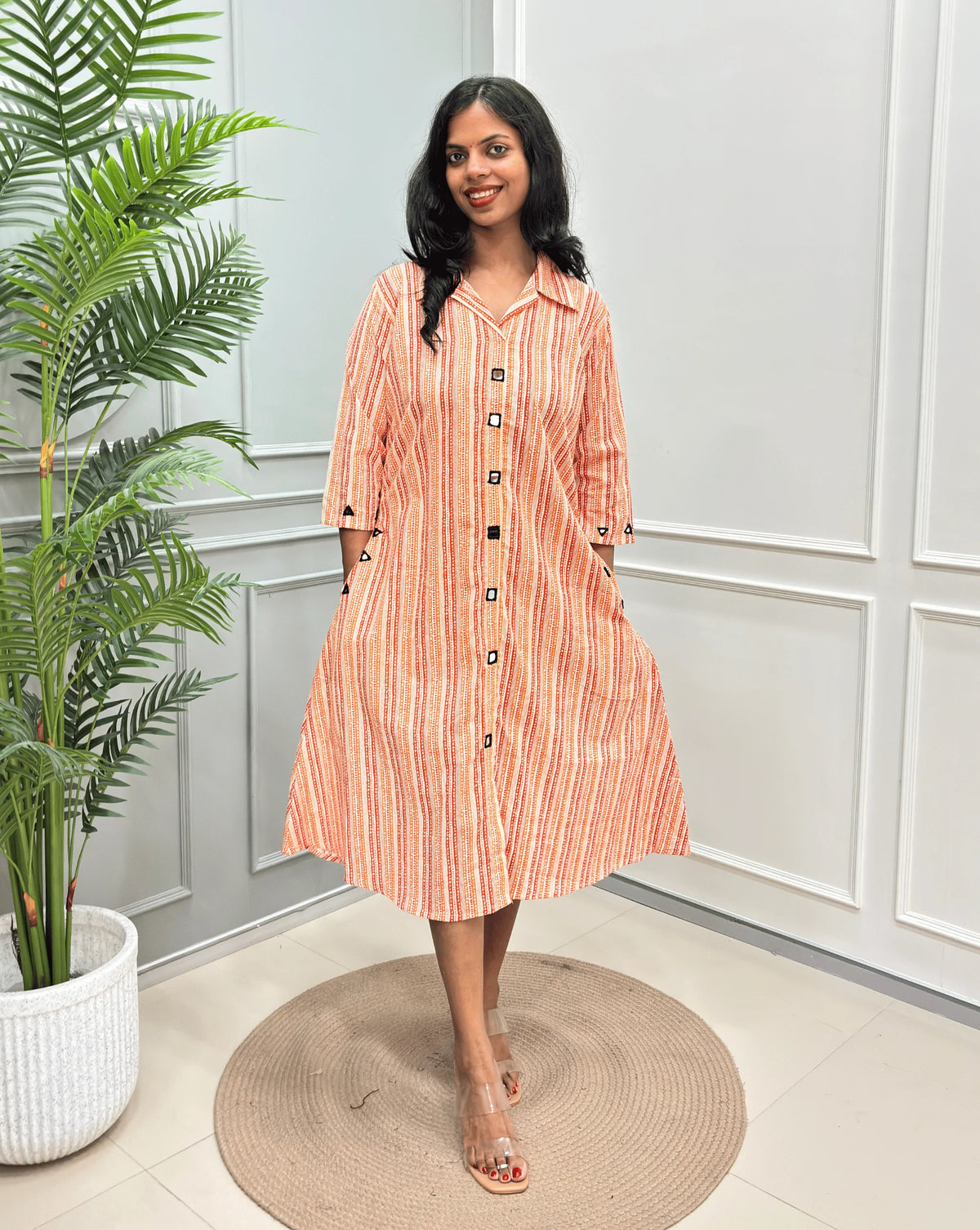 Rust Orange Women's Ajrakh A-line Pure Cotton Long Kurti with an Ajrakh Strip Print on abstract of orange, white, Rust orange,  standing on a round woven rug in front of a light gray paneled wall with a potted plant.