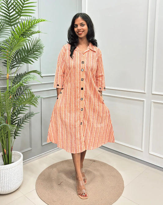 Rust Orange Women's Ajrakh A-line Pure Cotton Long Kurti with an Ajrakh Strip Print on abstract of orange, white, Rust orange,  standing on a round woven rug in front of a light gray paneled wall with a potted plant.