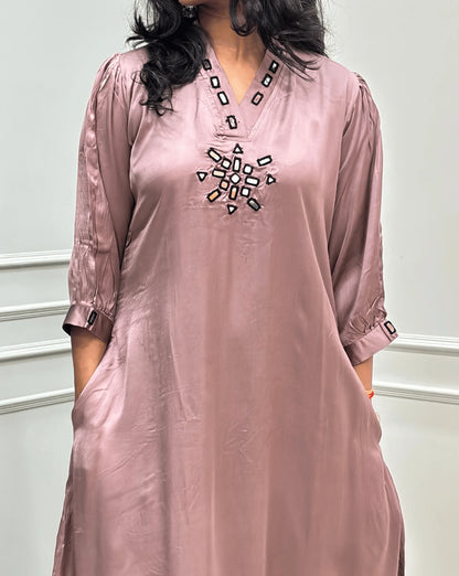 Close-up view of Onion Pink Women's Pure Modal Silk Ajrakh Long Kurti Featured with 3/4th Sleeves with a decorative Real Hand-Mirror embroidered V-neckline, Floral pattern on front & Sleeves Cuffs, standing on a round woven rug in front of a white paneled wall with a potted plant on the left