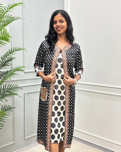 Front-View of Black women's Ajrakh pure Mashru silk Long one-peice V shape neck with beige border on Shrug & Three-quarter Sleeves, with Black Shrug attach with White inner consist floral print wiith black colour,  standing in a decorated room with a potted plant and paneled wall 