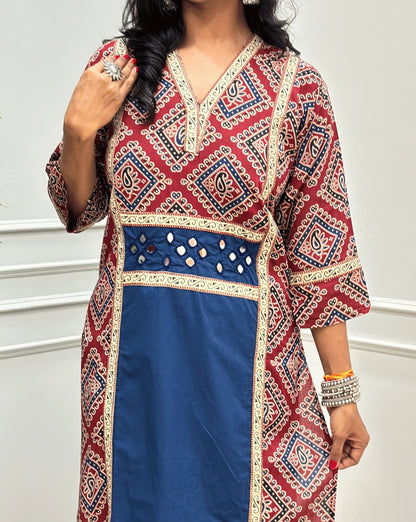 Close-up view of Blue Women's Ajrakh pure cotton 2 peice Co-Ord set featured with V Shaped neck with a Red base & an abstract of blue & Beige Bandhani print on Front Chest to Side & Sleeves, Hand-mirror work on Front as Belt, standing on a round woven mat in front of a white paneled wall with a potted plant.
