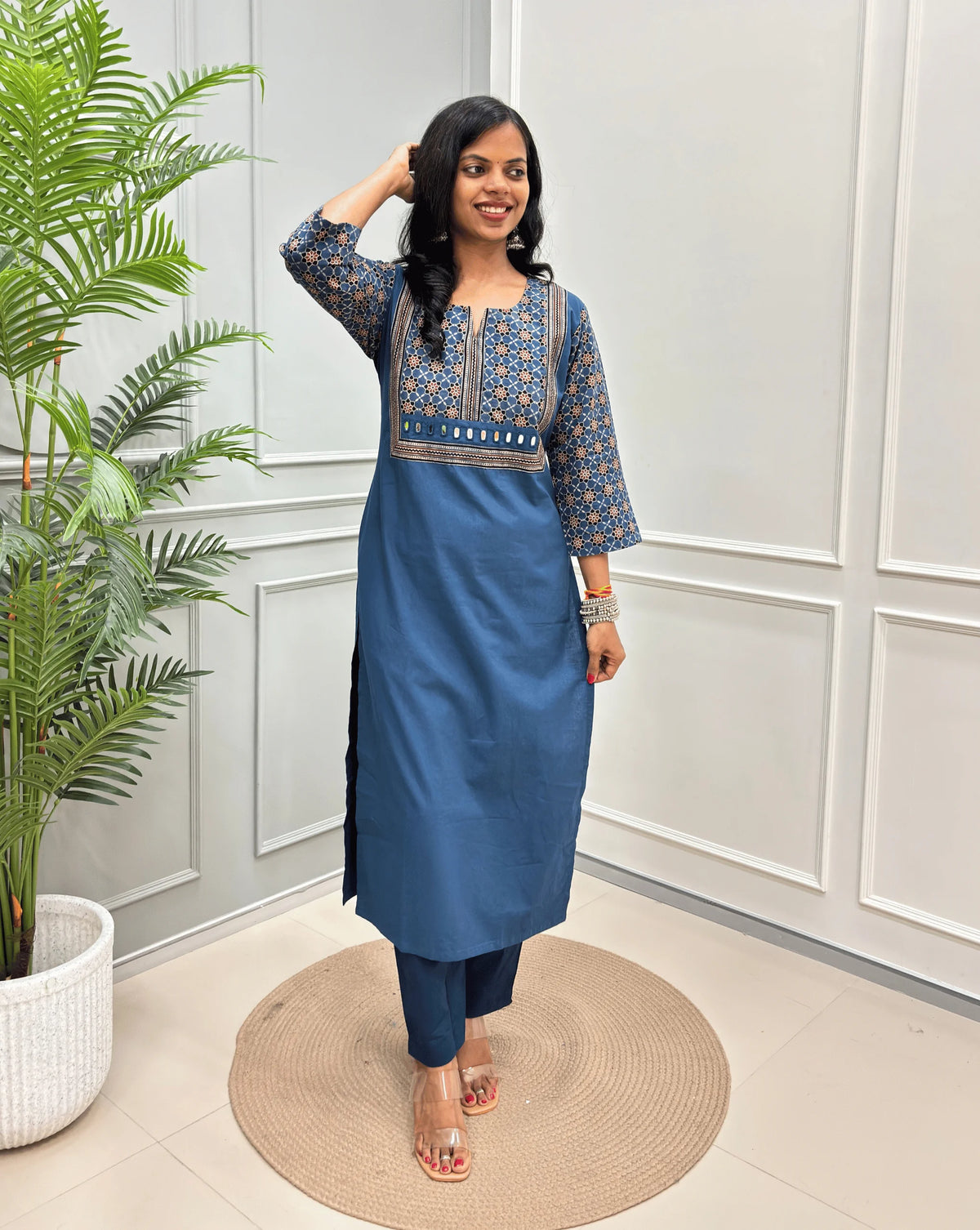 Light Blue Women's Hand-block Ajrakh Pure Cotton Long Kurti with a blue base , an abstract of black & beige Floral Print yoke on front & Full sleeves, paired with blue Pant, standing on a round woven mat in front of a white paneled wall.