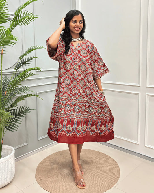Red Women's Ajrakh Pure Cotton loose-fitting kaftan Kurta with intricate white and beige geometric and floral patterns, standing on a round woven mat in front of a white paneled wall with a potted plant on the left.