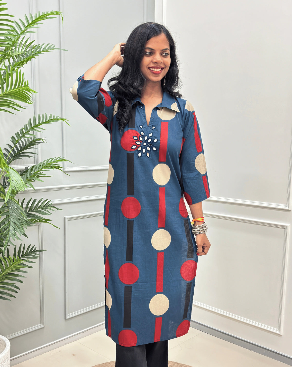 Front-view of dark Blue Women's Ajrakh Vegetable dyed plant based Cotton Long-Kurti with a geometric pattern of red, beige, and black circles, Hand-mirror work on front in flower shape & Sleeves cuffs,  paired with black Flared pant, standing in a room with a potted plant and paneled walls.
