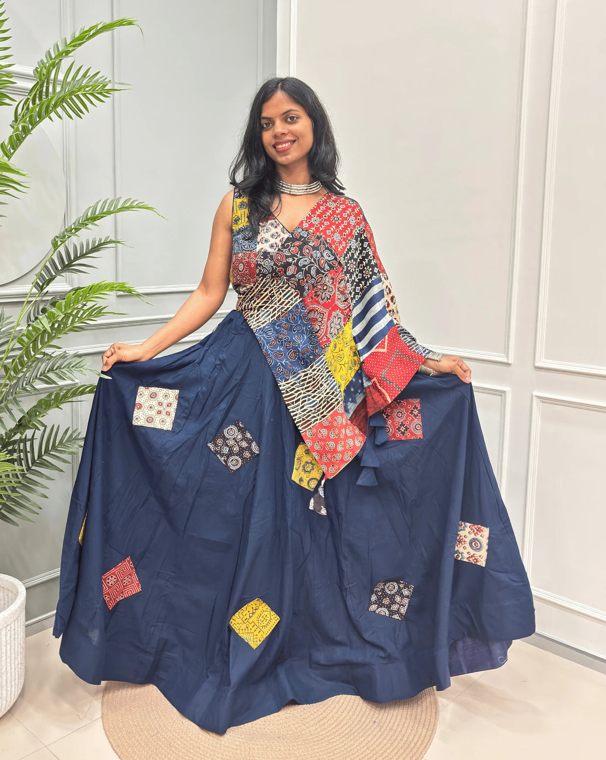 Navy blue Women's Ajrakh pure Cotton Skirt Set with a Ajrakh Patch work along with multi-colour Printed blouse & Dupatta, Standing against a light paneled wall with a green plant on the left.