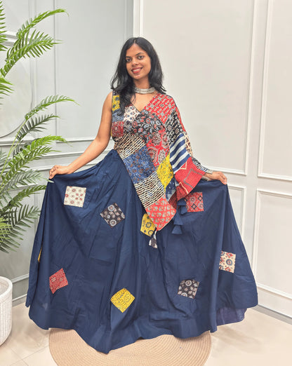 Navy blue Women's Ajrakh pure Cotton Skirt Set with a Ajrakh Patch work along with multi-colour Printed blouse & Dupatta, Standing against a light paneled wall with a green plant on the left.