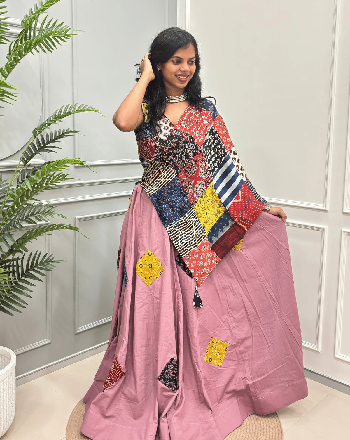 Front-View of Pink Women's Ajrakh pure Cotton Skirt Set with a beautiful Tasals & Ajrakh Patch work along with multi-colour Printed blouse & Dupatta, Standing against a light paneled wall with a green plant on the left.