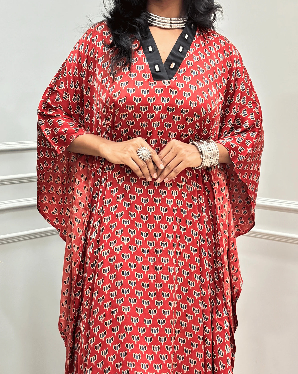 Close-up view of Red Women's Ajrakh Pure Modal Silk Knee Length Kaftan with a black Floral print, with Contrast Green Colour V-neck, along with Real Hand-Mirror embroidery, standing on a round woven rug in front of a white paneled wall next to a potted plant. 