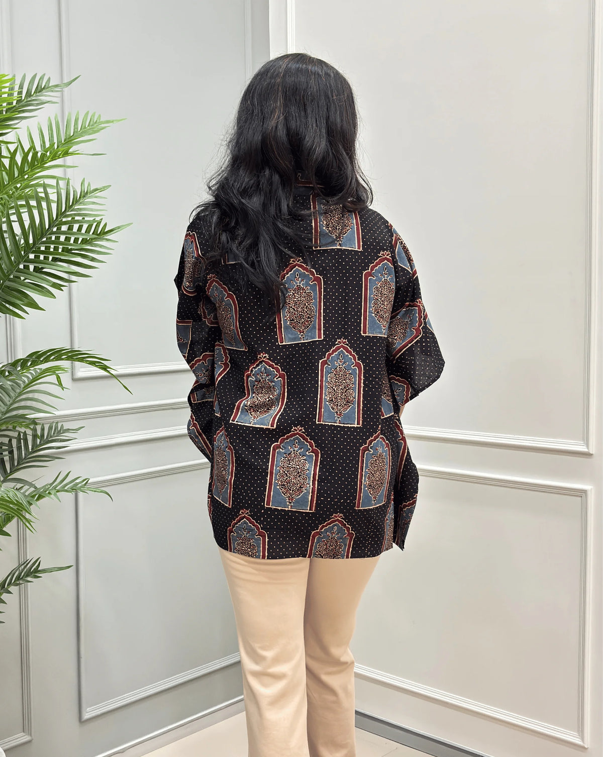 Close-up view of Black Women's Ajrakh Pure Cotton Short Kaftan Shirt Featured with Shirt Collar neck with a Grey, Red & Black Floral Block Print, Wooden Button on front , paired with beige flared pants, standing on a round woven rug in front of a white paneled wall with a potted plant.