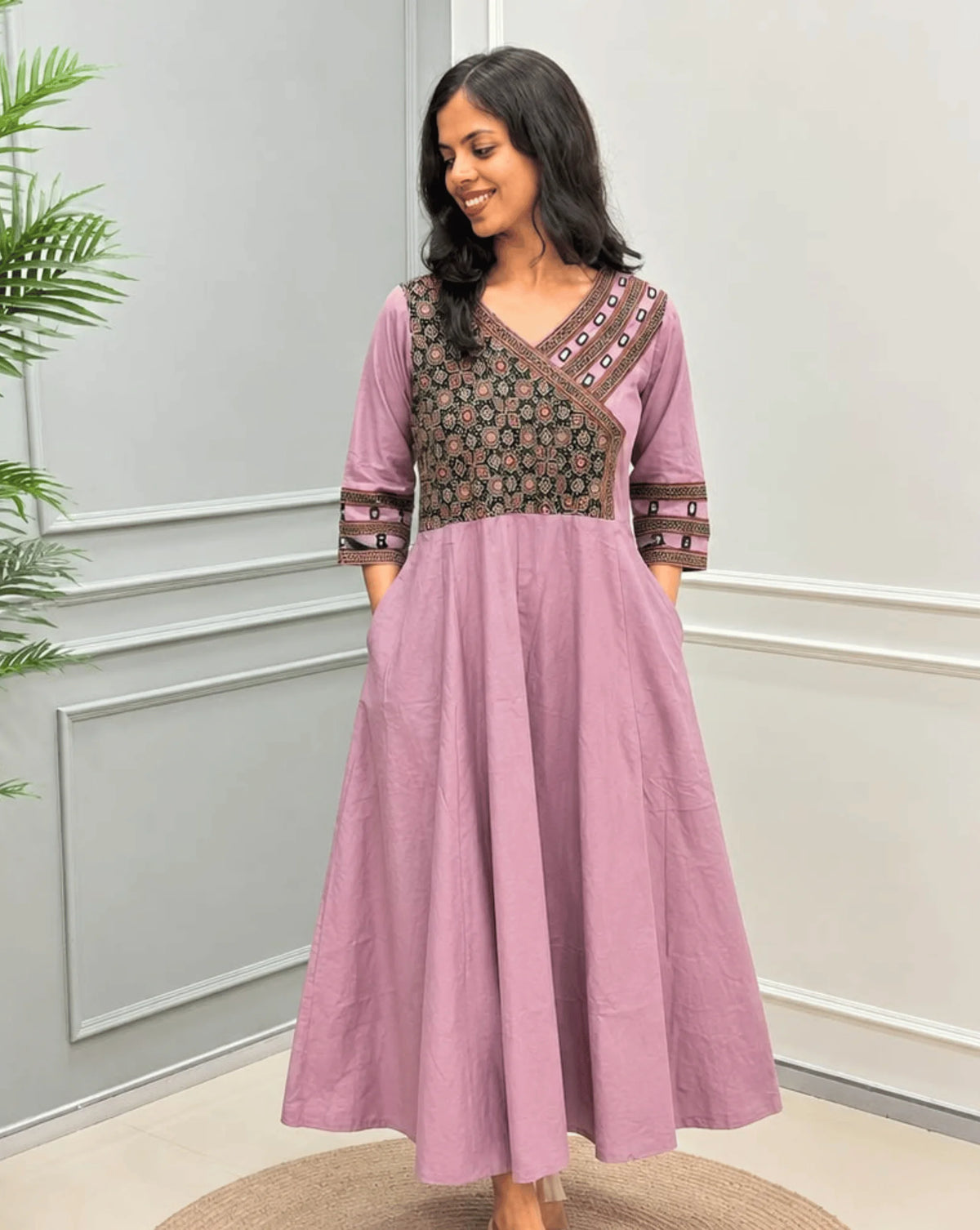 Front Veiw of Onion Pink  Women's Ajrakh Pure Cotton A-line Anarkali with a Black base & Red floral yoke patterned along Real Hand-mirror work on Front & sleeve cuffs,  standing on a round woven rug in front of a light gray paneled wall.