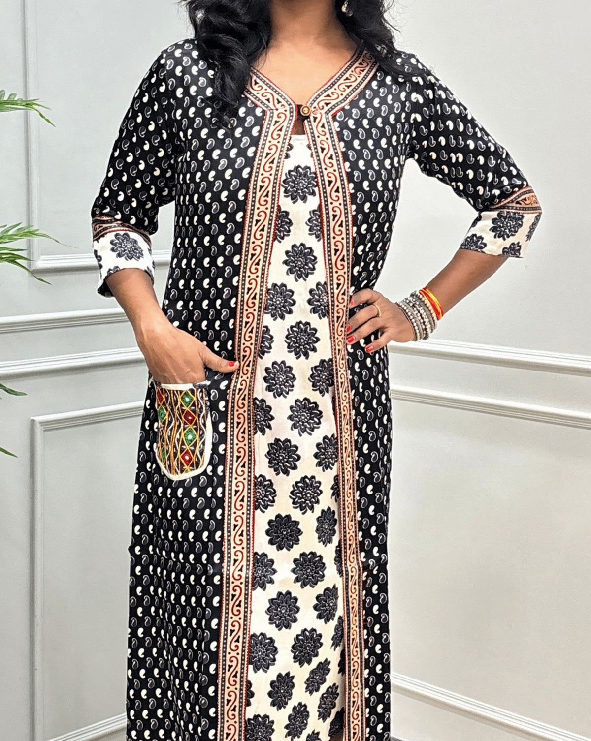Close-up of Black women's Ajrakh pure Mashru silk Long one-peice V shape neck with beige border on Shrug & Three-quarter Sleeves, with Black Shrug attach with White inner consist floral print wiith black colour, Kutchi thread embroidery on Front Side pocket,   standing in a decorated room with a potted plant and paneled wall 