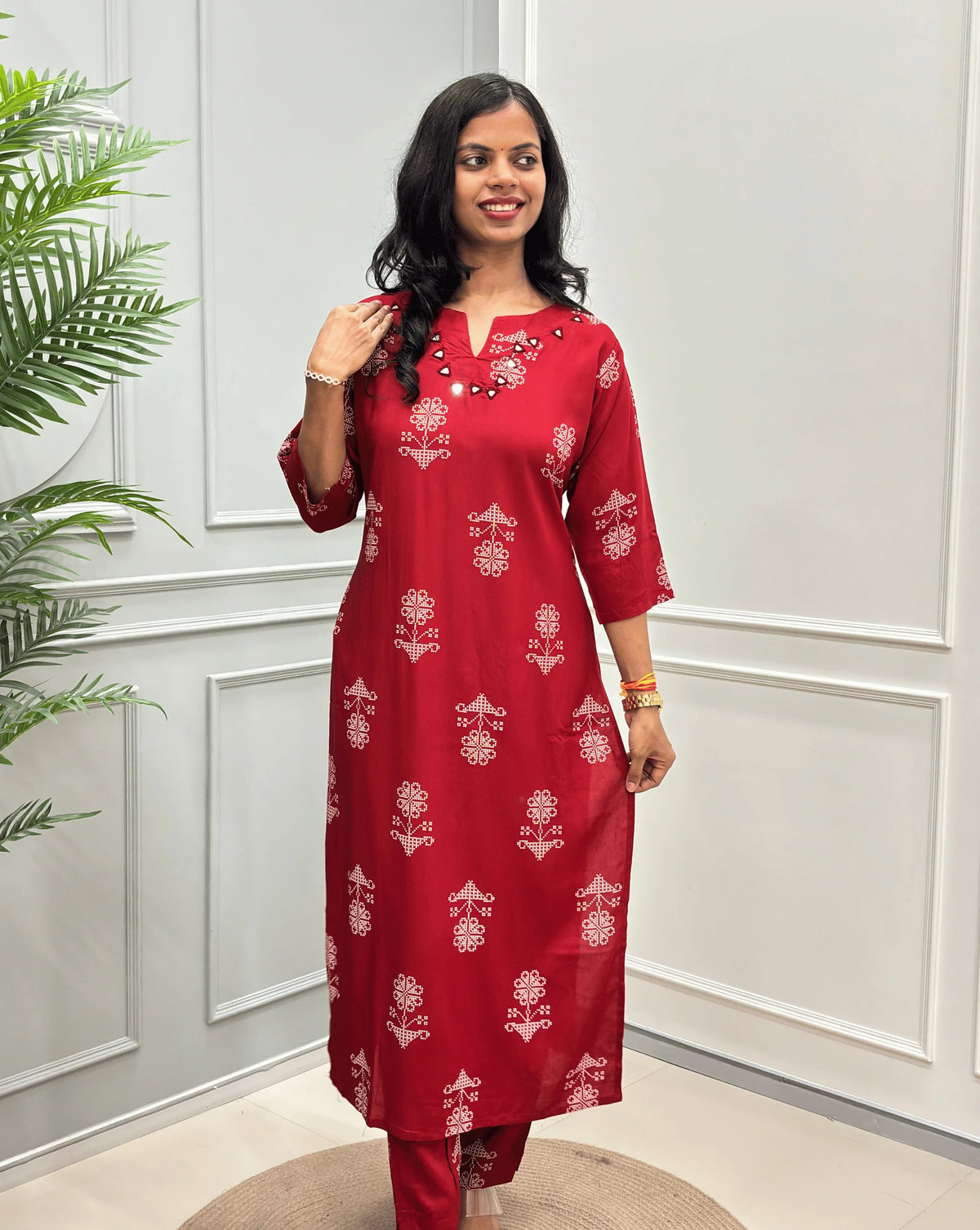 Front-view Red Women's Ajrakh Vegetable Dyed Cotton Long Co-Ord set with white floral prints & Real Hand-Mirror Work on neck-line, along with Same Matching pant,  standing on a round woven mat in front of a white paneled wall with a potted plant on the left.