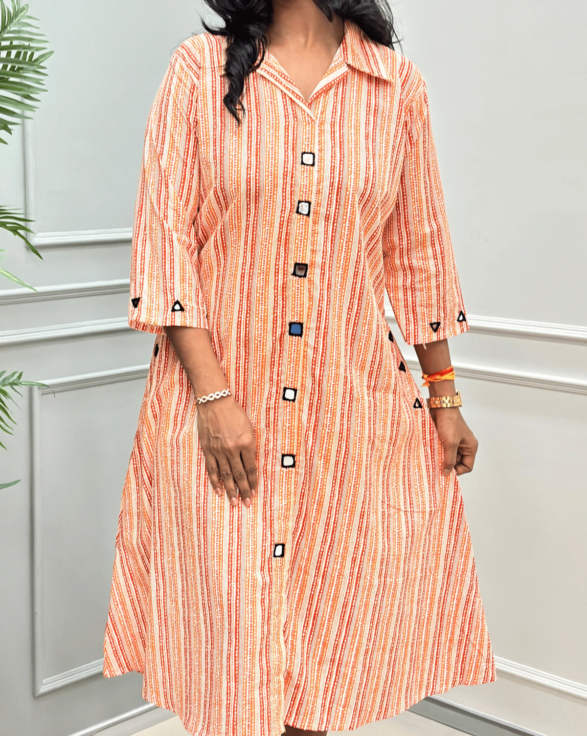 Close-up View of Rust Orange Women's Ajrakh A-line Pure Cotton Long Kurti Featured with Collar V neck & 3/4th Quarter Sleeves with an Ajrakh Strip Print on abstract of orange, white, Rust orange, Real hand-mirror work on front & Sleeves, Along with 2 side pockets, standing on a round woven rug in front of a light gray paneled wall with a potted plant.