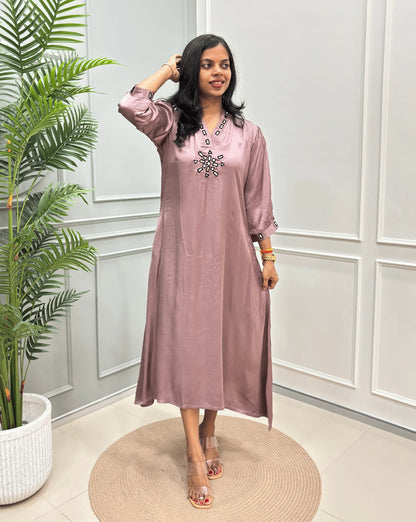 Onion Pink Women's Pure Modal Silk Ajrakh Long Kurti with a decorative Real Hand-Mirror embroidered neckline, Floral pattern on front & Sleeves Cuffs, standing on a round woven rug in front of a white paneled wall with a potted plant on the left