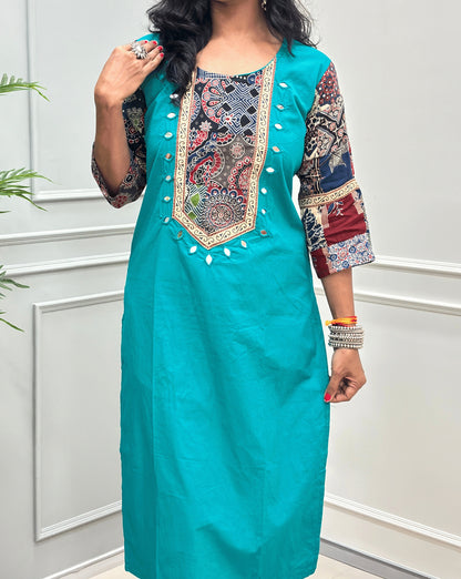 Close-up of Aqua Blue women's Ajrakh Vegetable dyed plant based Cotton Co-Ord set Featured with Round neck & Three-quarter sleeves with Multi-colour ajrakh Print detailed embroidered on neckline & Sleeves with beige strip border on work , Hand-mirror work out-line on front, paired with matching teal pants, standing on a round woven mat in a bright room with a potted plant.