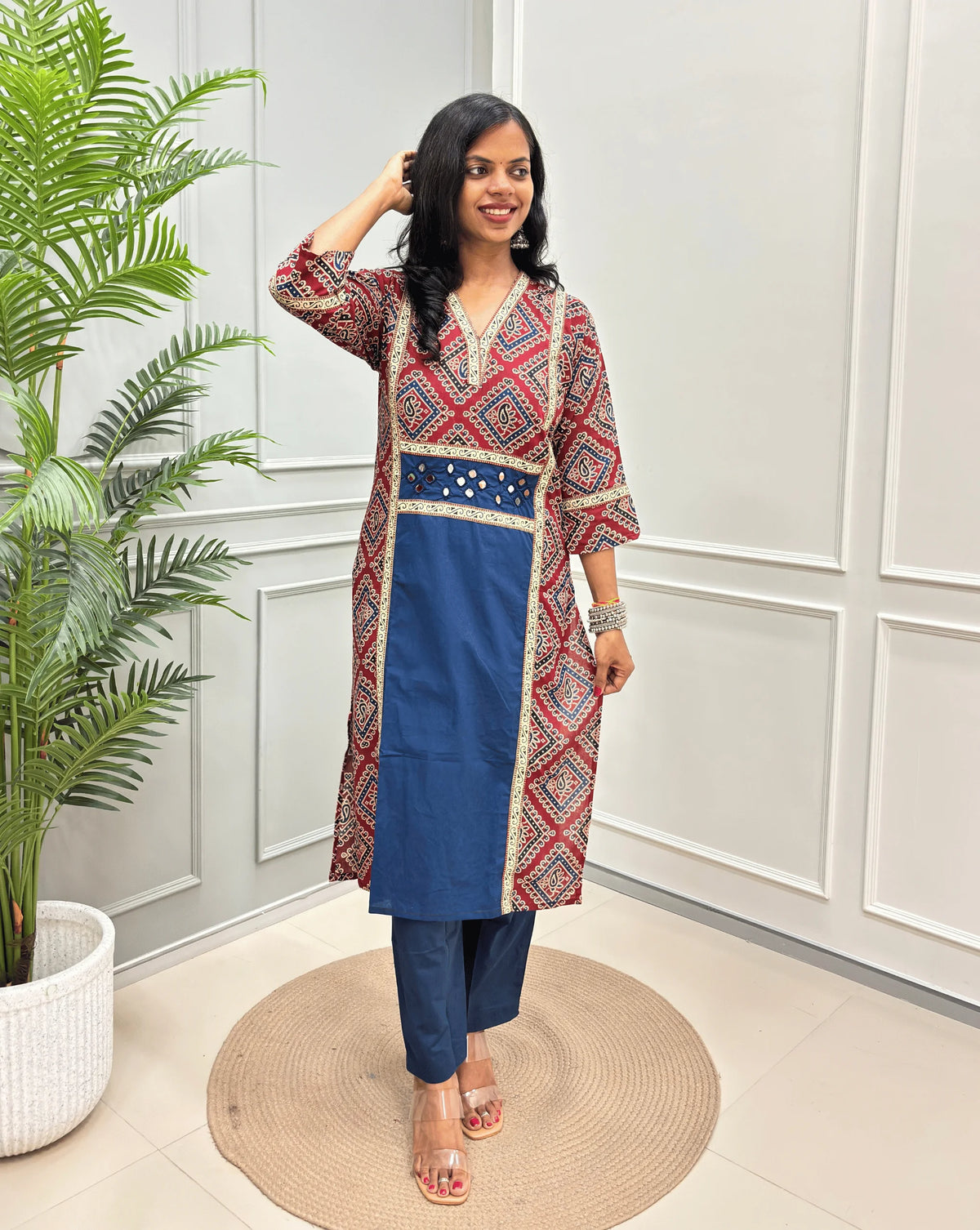 Blue Women's Ajrakh pure cotton 2 peice Co-Ord set with a Red base & an abstract of blue & Beige Bandhani print on Front Chest to Side & Sleeves , standing on a round woven mat in front of a white paneled wall with a potted plant.