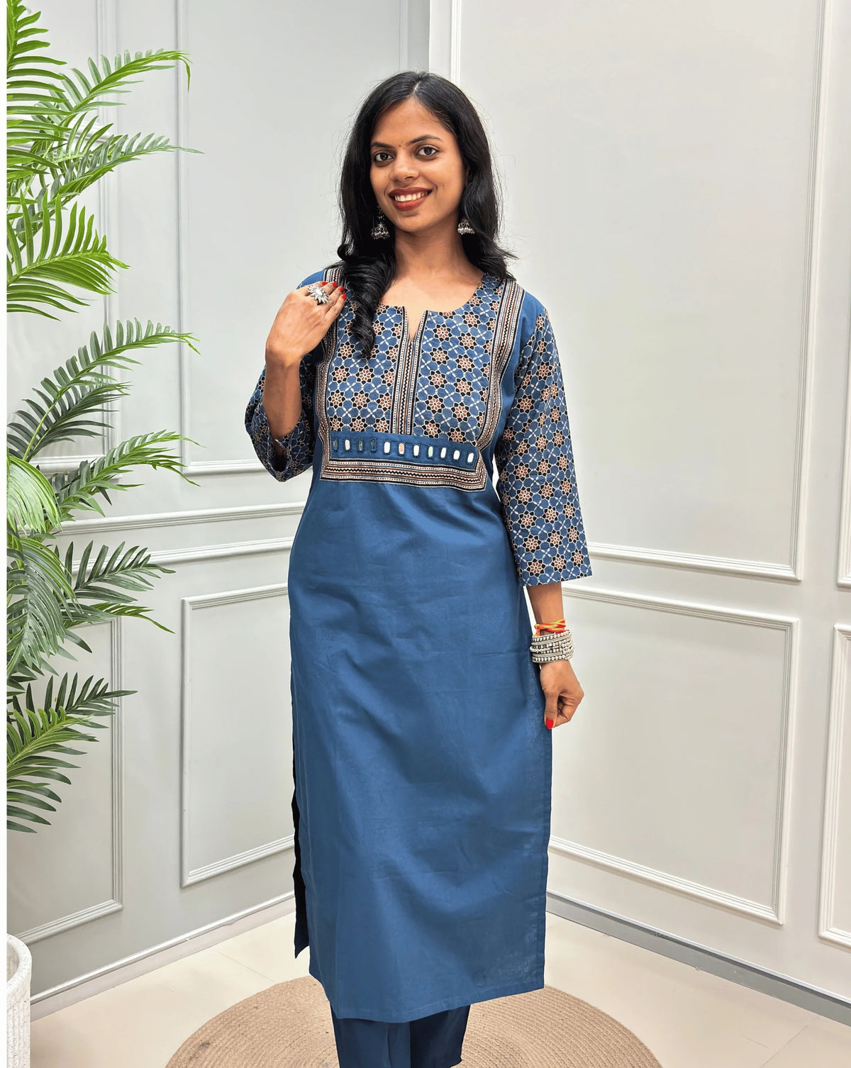 Front view of Light Blue Women's Hand-block Ajrakh Pure Cotton Long Kurti with a blue base , an abstract of black & beige Floral Print yoke on front & Full sleeves, Hand-mirror work on front, paired with blue Pant, standing on a round woven mat in front of a white paneled wall. 