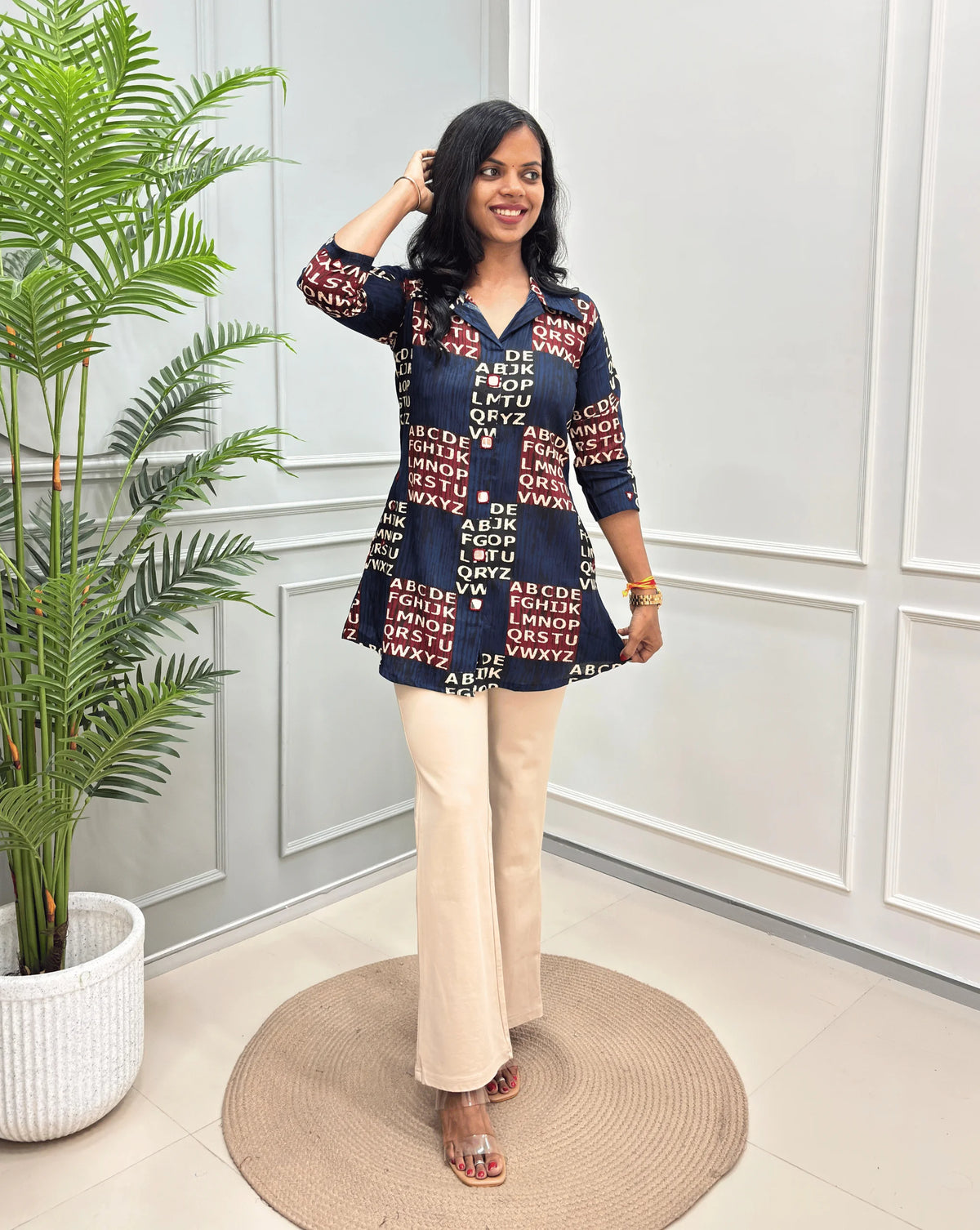 Navy Blue Women's Ajrakh Vegetable Dyed Plant based Cotton Short kurti with a Red & Black Base, & Abstract Beige color alphabet print, paired with beige trousers, standing on a round woven rug in front of a white paneled wall with a potted plant.