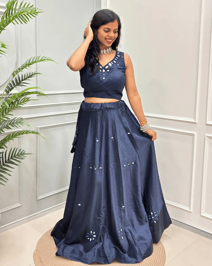 Navy-blue Women's Pure Mashru silk  two-piece traditional outfit consisting of a sleeveless crop top with Real Hand mirror work and a matching flared skirt with embroidered details, set against a white paneled wall with a green plant on the left.