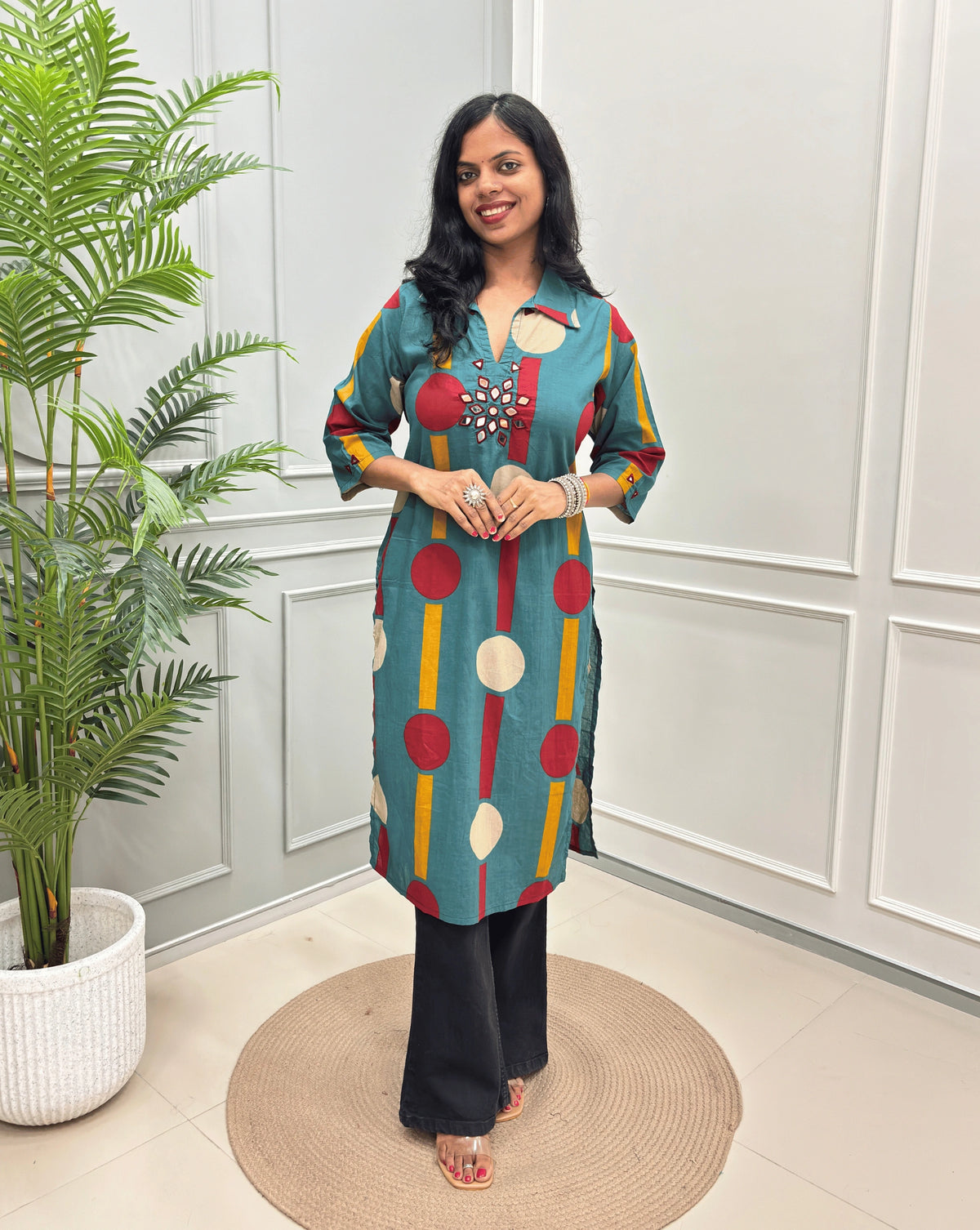 Light Green Women's Ajrakh Vegetable dyed plant based Cotton Long-Kurti with a geometric pattern of Red, white, and yellow circles, paired with black Flared pant, standing in a room with a potted plant and paneled walls.