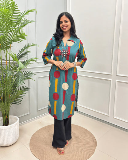Light Green Women's Ajrakh Vegetable dyed plant based Cotton Long-Kurti with a geometric pattern of Red, white, and yellow circles, paired with black Flared pant, standing in a room with a potted plant and paneled walls.