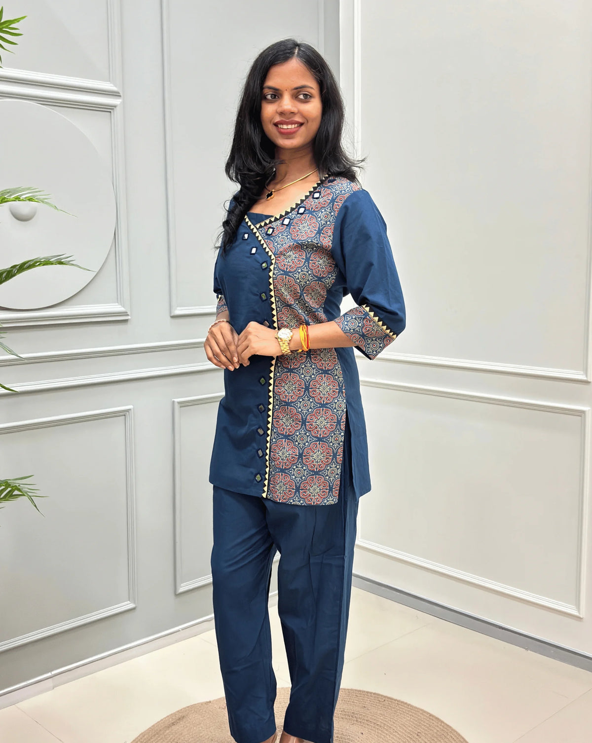 Front-view of Blue Women's Ajrakh Pure Cotton 2-peice Co-Ord set with a Ajrakh printed panel on the front & Sleeves, Angrakha Patterned on front, Real Hand-Mirror work on front Along with 2 side pockets in pant, standing on a round woven rug in a modern room with white paneled walls.