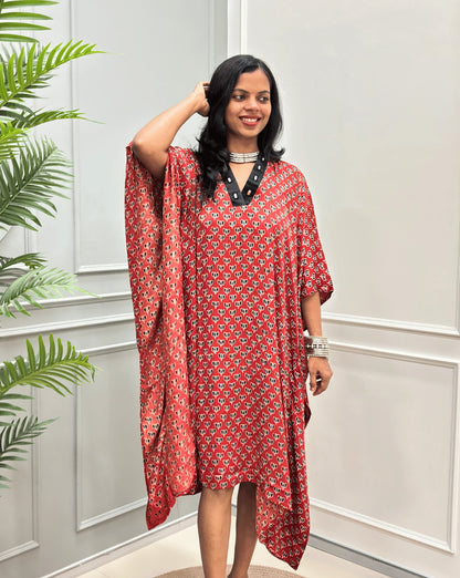 Front-view of Red Women's Ajrakh Pure Modal Silk Knee Length Kaftan with a black Floral print, with Contrast Black Colour V-neck, standing on a round woven rug in front of a white paneled wall next to a potted plant.