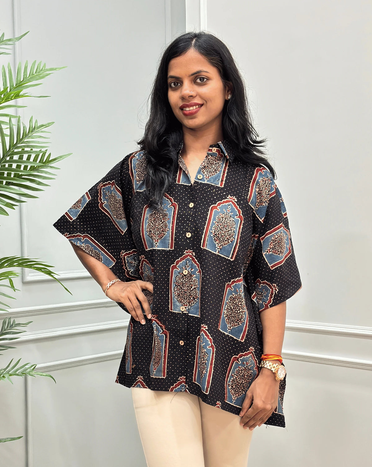 Front-view of Black Women's Ajrakh Pure Cotton Short Kaftan Shirt with a Grey, Red & Black Floral Block Print, Wooden Button on front , paired with beige flared pants, standing on a round woven rug in front of a white paneled wall with a potted plant.
