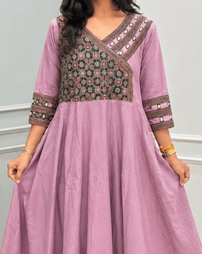 Close-up Veiw of Onion Pink Women's Ajrakh Pure Cotton A-line Anarkali Featured with V-neck with a Black base & Red floral yoke patterned along Real Hand-mirror work on Front & sleeve cuffs , 2-Side pockets, standing on a round woven rug in front of a light gray paneled wall.
