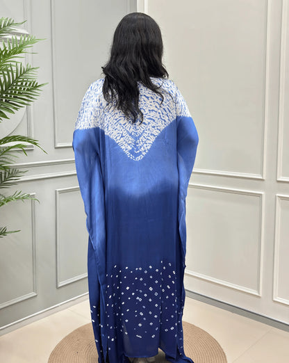 Back-Side View of Blue Women's Ajrakh Pure Modal Silk Shibori Kaftan Featured with Long-length, with a 3 shades of Blue with ajrakh print on chest & Bandhani Small dots with white on Bottom, standing on a round woven rug in front of a white paneled wall.