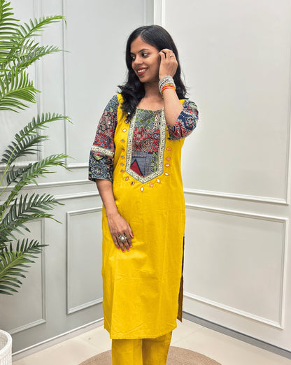 Front-view of Yellow women's Ajrakh Vegetable dyed plant based Cotton Co-Ord set with Multi-colour ajrakh Print detailed embroidered on neckline & Sleeves with beige strip border on work , Hand-mirror work out-line on front, paired with matching teal pants, standing on a round woven mat in a bright room with a potted plant.