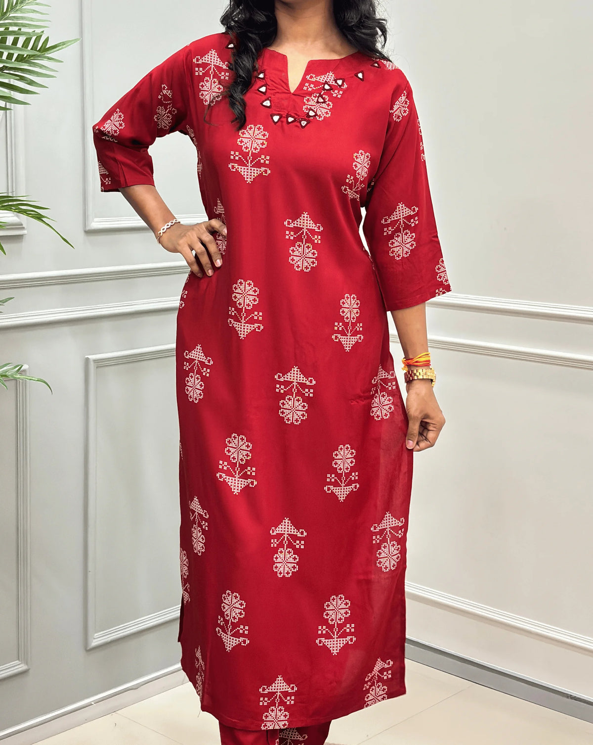Close-up of Red Women's Ajrakh Vegetable Dyed Cotton Long Co-Ord set Featured with Round V-neck & Three-Quarter Sleeves with white floral prints & Real Hand-Mirror Work on neck-line, along with Same Matching pant,  standing on a round woven mat in front of a white paneled wall with a potted plant on the left