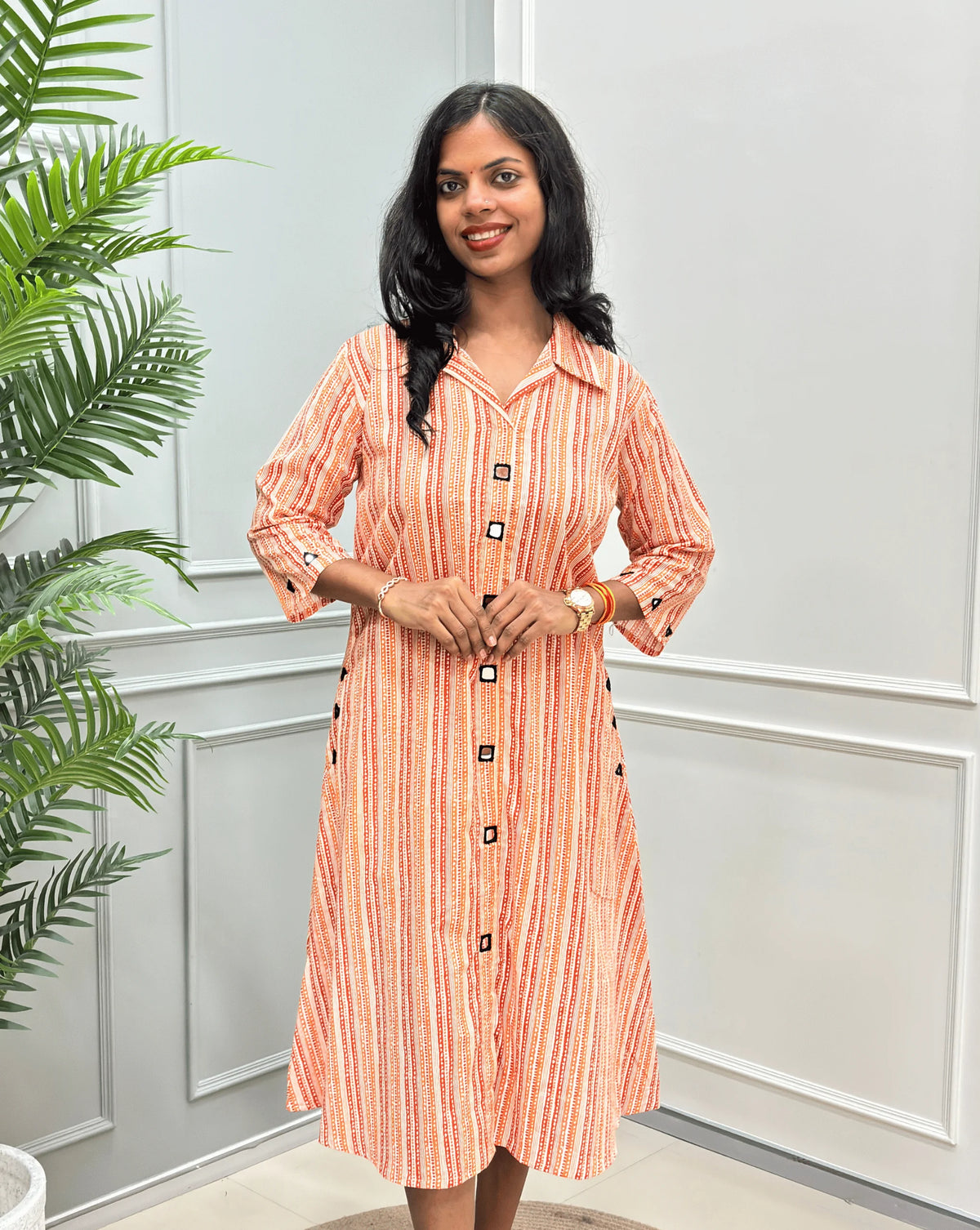 Front-View of Rust Orange Women's Ajrakh A-line Pure Cotton Long Kurti with an Ajrakh Strip Print on abstract of orange, white, Rust orange, Real hand-mirror work on front & Sleeves, Along with 2 side pockets, standing on a round woven rug in front of a light gray paneled wall with a potted plant.